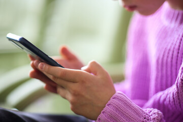 Teenage girl in a bright sweater sits on a green chair with headphones posting in social media apps, chatting with friends online on smartphone