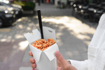 Woman holding paper box of takeaway noodles with fork outdoors, closeup. Street food