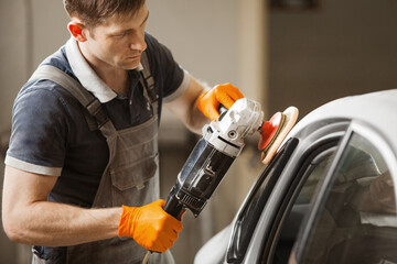 Car detailing - male worker holds a polisher in the hand and polishes white car in auto repair shop.