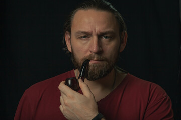 Lumberjack brutal beard muscled man in red shirt with smoking tube pipe standing on dark background