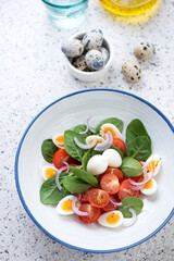 Plate with spinach, cherry tomatoes and quail eggs salad, vertical shot on a light-grey granite background, elevated view