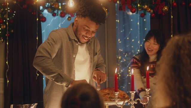 African American man cuts turkey or chicken with knife. Multi cultural friends celebrating Christmas or New Year. Served holiday table with candles. Warm atmosphere of Christmas dinner at home.