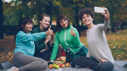 Attractive young ladies yogini are taking selfie using smartphone during picnic in park in autumn. Girls are posing and smiling having fun sitting together on mats.