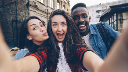 Point of view shot of joyful girls and guys multiethnic group taking selfie holding camera and posing outdoors during enjoyable vacation in beautiful city. Tourism and photography concept.