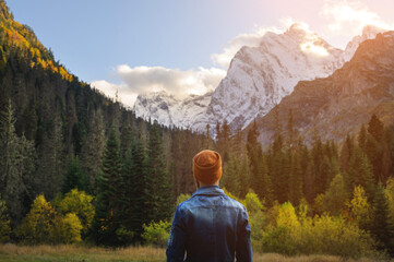 Back view of a man in a knitted hat and denim jacket against the backdrop of a green dense forest and snow-capped high mountains
