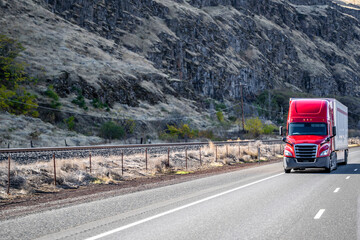 Bright red big rig long hauler semi truck with dry van semi trailer running on the road along the rocky mountain range