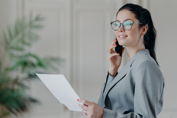 Photo of elegant brunette woman in formal clothes works with papers in office, discusses business issues via smartphone with colleague. Female manager checks information from documents, makes research