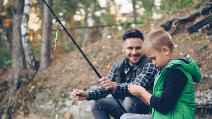 Adorable little boy is fishing with dad in pond in forest holding rod while his dad is talking to him explaining how to use gear. Family, hobby and wild nature concept.