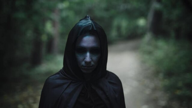 Portrait of a witch in a black cloak with a hood. Woman in the mystical forest. Dark forces