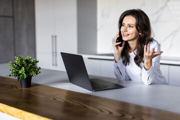 Photo of a young cheerful woman indoors at home at the kitchen using laptop computer talking by mobile phone.