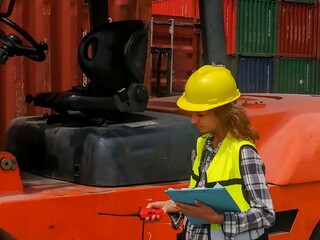 A female worker in a helmet and uniform is checking the quality of forklifts at a freight container...