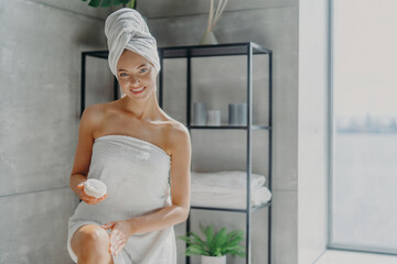 Smiling young European woman uses beauty prodcut for healthy glowing skin, applies cream on legs after taking shower, undergoes hygienic procedures at home poses in bathroom. Daily skin care procedure