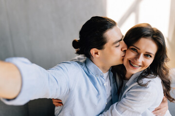 Young couple relaxing on a couch at home, hugging, taking a selfie