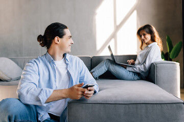 Young man and woman sitting together on couch while using laptop and smartphone