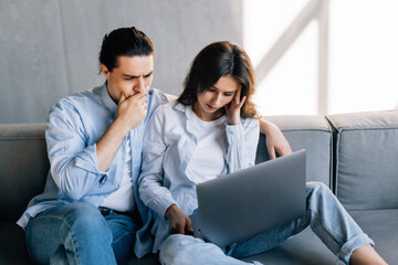 Confused upset married couple sit on couch read financial papers invoices troubled with too high expenses utility costs.