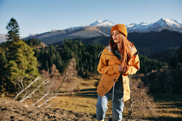 Woman hiking in the mountains in the fall with a smile with teeth and happiness in a yellow cape with red hair full-length stands against the backdrop of trees and mountains in the sunset light