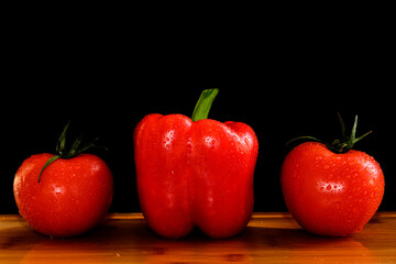 Two fresh red tomatoes and a paprika pepper with drops on a wooden table in a row. Black background.