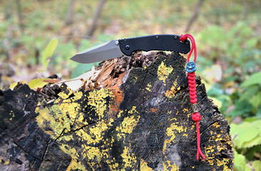 Folding knife cutting stainless steel blade black handle red lanyard blue bead green yellow leaves brown wood macro background