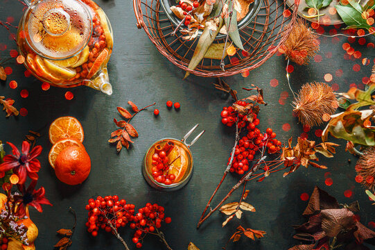 Autumn vitamin tea with Rowan berries, cinnamon sticks, orange and lemon slices in cup and teapot on dark table with ingredients, fall leaves and bokeh. Cozy seasonal composition. Healthy lifestyle