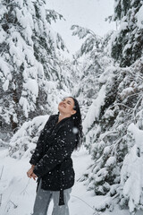 A happy girl is walking in a snowy forest. Snowfall. The girl has dark long hair. The woman is wearing a black jacket and light jeans. Christmas trees are fastened around her.