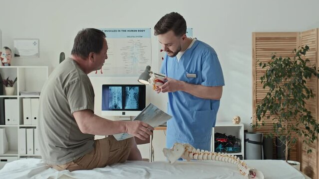 Therapist in uniform explaining the problem with spine to patient showing the model of skeleton during medical exam at hospital