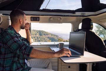 young man working on his laptop from his camper van in the middle of the nature, concept of freedom and digital nomad lifestyle
