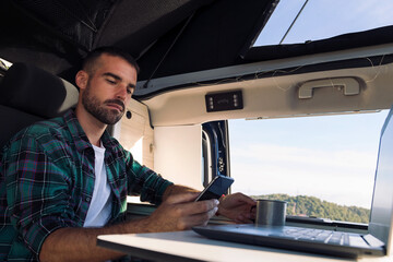 young man using his mobile phone and laptop from his camper van in the middle of nature, concept of...