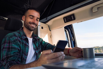 smiling young man using his mobile phone and laptop from his camper van in the middle of nature,...