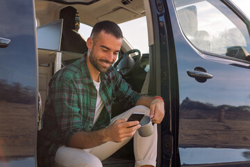 smiling young man using his mobile phone sitting on the door of his camper van, concept of freedom...