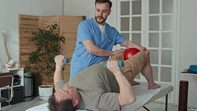 Physiotherapist in uniform showing exercises with dumbbells to mature patient while he lying on couch