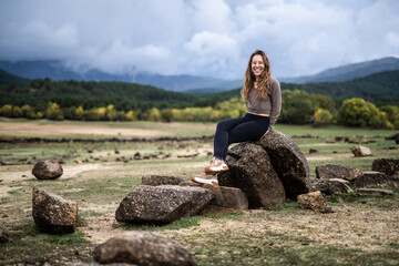 Naklejka na ściany i meble Young beautiful girl sitting on a rock in the middle of a lake with a mountain landscape background smilling at the camera in winter season