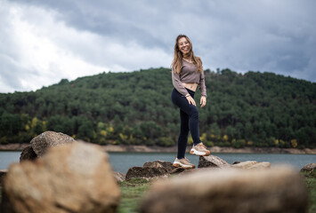 Naklejka na ściany i meble Young beautiful girl on a rock in the middle of a lake with a mountain landscape background smilling at the camera in winter season