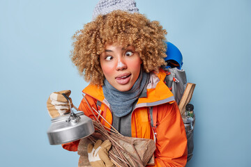 Horizontal shot of funny curly female camper makes grimace at camera carries wood and kettle...
