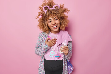 Happy young curly expectant mother enjoys pregnancy time smiles broadly expresses sincere emotions prepares for motherhood poses with baby items isolated over pink background dreams about child