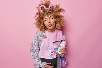 Affectionate future mother keeps lips rounded touches belly being on third trimester holds feeding bottle carries net bag sends air kiss at camera isolated over pink background. Expectancy concept