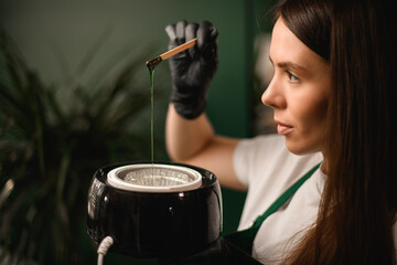 beautician accurate holds heating device and wooden stick with flowing green wax for depilation.