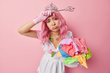 Obraz na płótnie Canvas Unhappy tired housewife poses with basin full of colorful laundry wears crown and white dress holds magic wand pretends being fairy of cleanliness isolated over pink background does housework