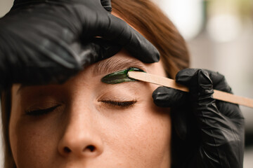 Close-up of the process of applying green wax to skin at female eyebrows - obrazy, fototapety, plakaty