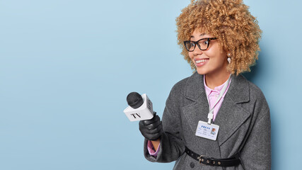 Public speak television and broadcasting concept. Positive curly haired woman correspondent holds microphone wears elegant clothes isolated over blue background copy space for your advertisement.