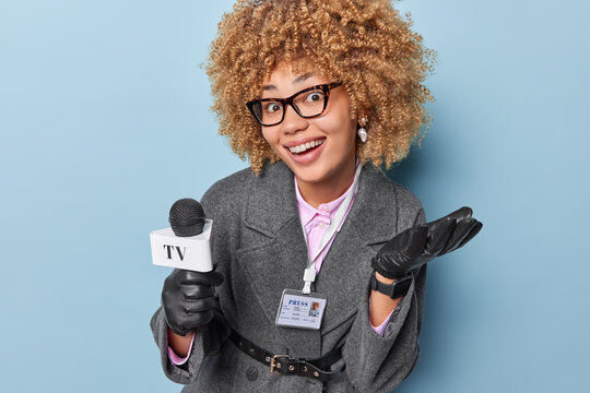 Positive curly haired female TV presenter holds microphone prepares reportage tells news on her channel wears spectacles grey coat and rubber gloves isolated over blue background. Television concept
