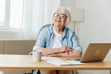 Fototapeta na wymiar a happy elderly woman with gray hair is sitting at her desk with a laptop and smiling broadly looks away with her hands folded on the table. The concept of working from home
