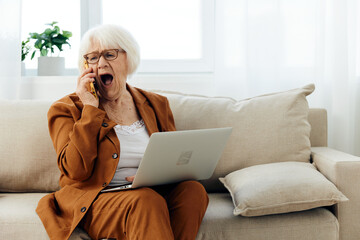 a sad, elderly businesswoman is sitting on a sofa in a cozy apartment in a brown suit working from...