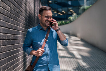 Cheerful handsome young adult man, making a phone call, standing near a wall.