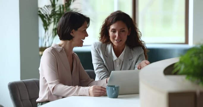 Hispanic millennial woman and middle-aged female colleagues working together meet in office staring at laptop screen, learn new software, younger mate helps to older with business app use modern tech