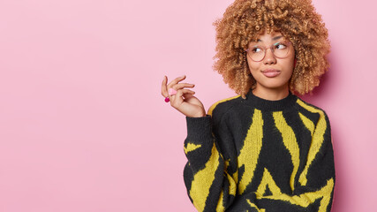 Doubtful pensive woman with curly hair keeps hand raised shrugs shoulders focused aside feels confused wears black and yellow jumper spectacles for vision correction stands against pink background