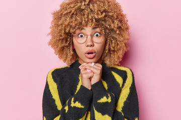 Portrait of stunned woman with curly blonde hair keeps mouth opened gasps from wonder keeps hands under chin wears big transparent eyeglasses and casual jumper isolated over pink background.