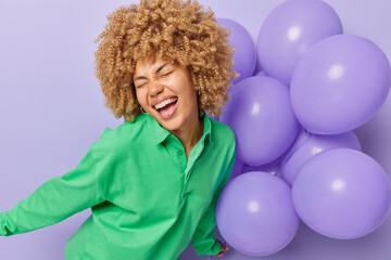 Cheerful overjoyed young woman with curly hair enjoys birthday holiday party laughs out gladfully dressed in green shirt holds bunch of inflated helium balloons isolated over purple background.