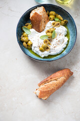 Blue bowl with ricotta, green olives, olive oil and crusty bread on a beige stone background, vertical shot with space, high angle view