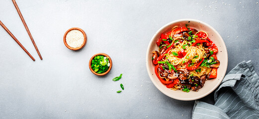 Vegetarian stir fry noodles with vegetables, paprika, mushrooms, chives and sesame seeds in bowl. Gray table background, top view, copy space