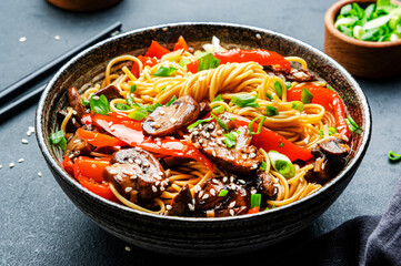 Vegan stir fry egg noodles with vegetables, paprika, mushrooms, chives and sesame seeds in bowl. Asian cuisine dish. Black table background, top view